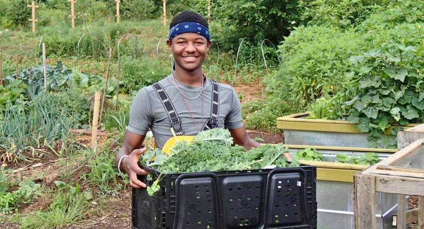 A person smiles at the camera while carrying a box filled with vegetables. A garden is in the background.